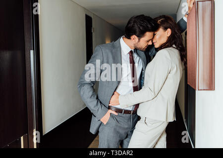 handsome man in suit standing with hand in pocket and kissing attractive woman in hotel corridor Stock Photo