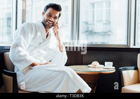 cheerful bearded man in white bathrobe sitting on chair and talking on smartphone Stock Photo