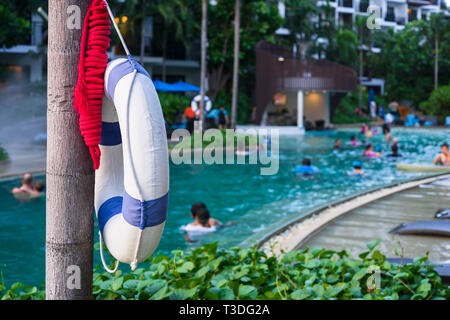 Lifebuoy hanging on a tree next to the outdoor swimming pool for the safety of people with blurred people swimming on a swimming pool. Stock Photo