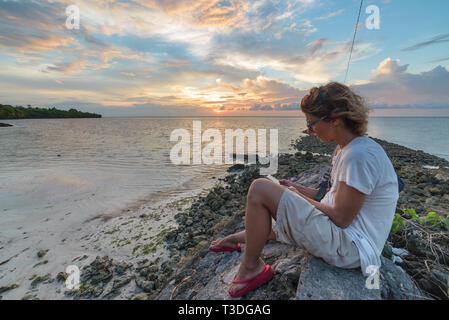 Adult woman with eyeglasses reading ebook relaxing at tropical beach dramatic sky holidays vacation concept Stock Photo