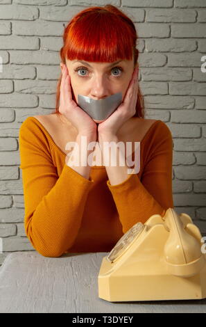 Conceptual picture. Young red-haired girl sitting near an old dial-up phone with adhesive tape lips to keep silence Stock Photo