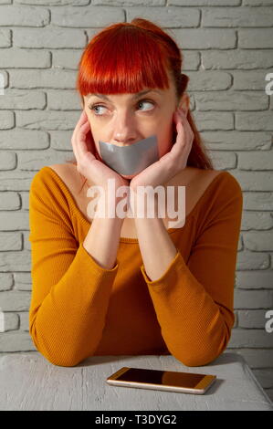 Conceptual picture. Young red-haired girl sitting near a smartphone with adhesive tape lips to keep silence Stock Photo