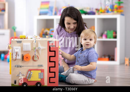 Cute toddler baby playing with busyboard. Mother or teaching kid in nursery. Children's educational toys. Stock Photo