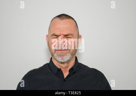 Upset bereft man with closed eyes in anguish with a pained expression, head and shoulders portrait over a grey studio background Stock Photo