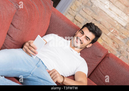 A happy handsome young man in a white tshirt laying on a sofa and using his smartphone. Stock Photo