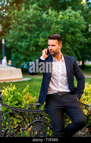 A handsome young businessman leaning against the fence in a park while talking on a phone Stock Photo