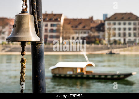 A ship's bell hangs from a mooring post on the bank of the Rhine in Basel, Switzerland. Passengers ring it to summon the ferry when the boat waits on the far shore. In the background, the wooden reaction ferry "Leu" crosses the river between Kleinbasel and Grossbasel, pulled only by the current along a taut steel cable. Stock Photo