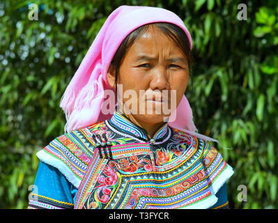 Mature Miao woman (Chinese ethnic minority) wears a colourful traditional ethnic costume with a pink headscarf and poses for the camera. Stock Photo