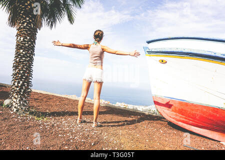 Back view of a young woman raising her hands up while admiring the panoramic view of ocean from the hill - Beautiful girl having fun traveling Stock Photo