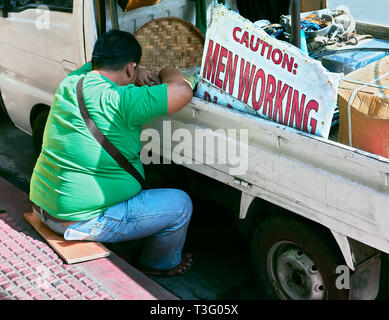 Manila, Philippines: Overweight man sitting on the floor leaning against a car sleeping, next to signage 'Caution Men Working' Stock Photo