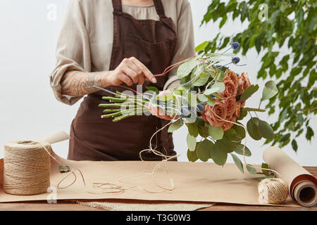 Floral design studio, woman's hands with tatto are making decorative bouquet from fresh natural fragrant flowers roses living coral color on a gray ba Stock Photo