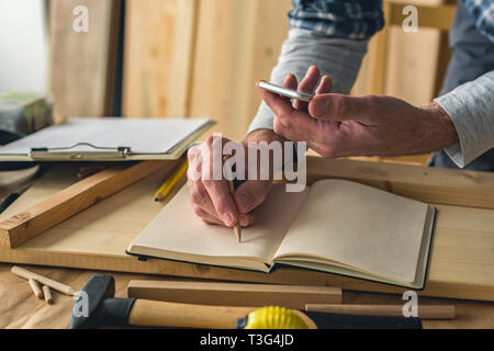 Carpenter is writing project notes while using smartphone in small business woodwork workshop Stock Photo