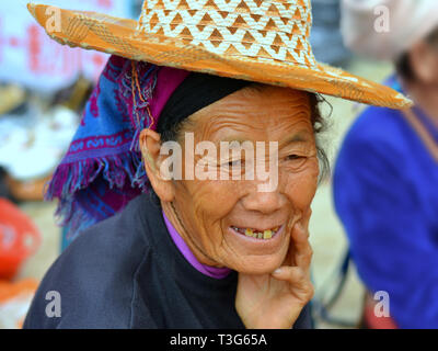 Old Hani market woman (Chinese ethnic minority) with lived-in face wears a straw hat over her blue tribal headscarf. Stock Photo