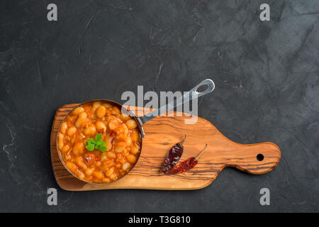 Baked beans in tomato sauce served in pan with basil and chili pepper on dark background with copy space. Flat lay Stock Photo