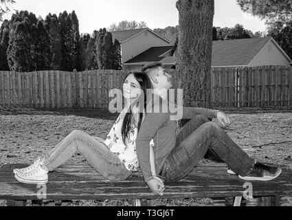 Caucasian couple in love relaxing while sitting on top of picnic able in backyard with their backs to each other.  Black and white image. Stock Photo