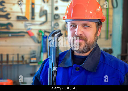 A bearded white middle-aged man in a helmet and with a big key in his hand is looking directly into the camera. Portrait of a plumber. Stock Photo