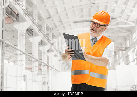 The builder in a construction vest and orange helmet standing against industrial background. Safety specialist, engineer, industry, architecture, manager, occupation, businessman, job concept Stock Photo