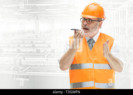The builder in a construction vest and an orange helmet holding smartphone, using voice control against industrial background. Safety specialist, engineer, industry, architecture, manager, occupation, businessman, job concept Stock Photo