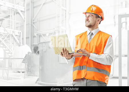 The builder in a construction vest and orange helmet smiling with laptop against industrial background. Safety specialist, engineer, industry, architecture, manager, occupation, businessman, job concept Stock Photo