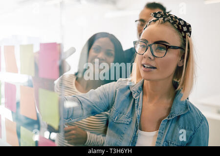 Young businesswoman and her team brainstorming together with sticky notes on the glass wall of a modern office Stock Photo