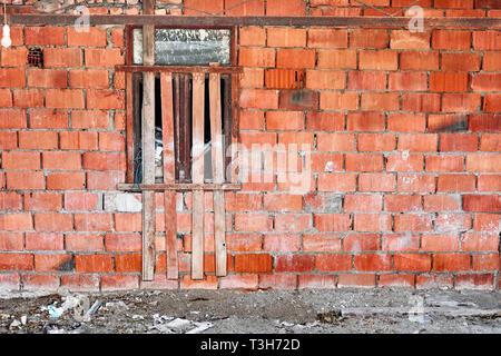 Red brick wall and window of an abandoned unfinished building in a construction site. The window is reinforced and secured with wooden plank bars. Stock Photo