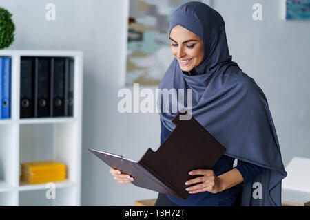 Beautiful Arabian woman wearing headscarf and reading documents at the office. Radiant smile and satisfied by work. Stock Photo
