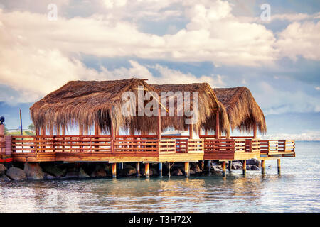 Three beach huts with palm leaf covered roofs on a wooden platdorm over the sea at sundown. Beautiful seascape and cloudscape view. Stock Photo