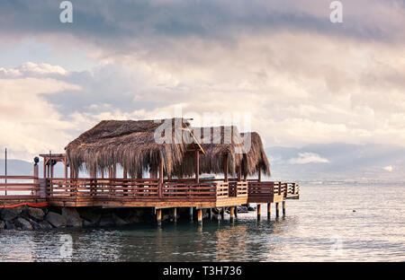 Three beach huts with palm leaf covered roofs on a wooden platdorm over the sea at sundown. Beautiful seascape and cloudscape view. Stock Photo