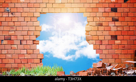 Broken red  brick wall with a big hole in the middle and blue sky, clouds and sun view through the hole. Stock Photo