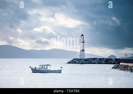 Lighthouse and white fishing boat over the calm sea on a cloudy winter morning in Turgutreis harbor, Bodrum, Turkey. Stock Photo