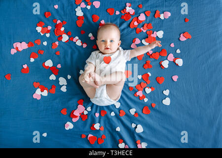 Cute adorable white Caucasian baby girl boy infant with blue eyes four months old lying on bed among many foam paper red pink colorful hearts. View fr Stock Photo