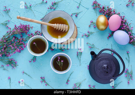 Tea with honey, easter eggs and purple heather flowers on blue background. Flat lay. Stock Photo