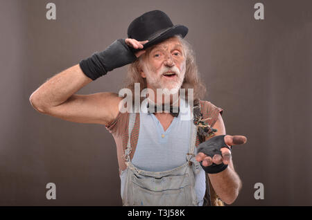 Old and dirty homeless beggar touching his black hat and asking for money, studio shot Stock Photo