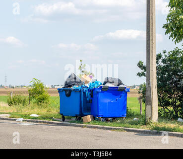 Overflowing blue plastic waste cans with black garbage bags, cardboard boxes and other garbage outside the city near the road. One waste can is broken Stock Photo