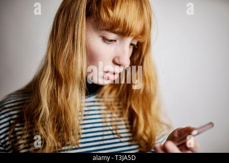 Girl, teenager, red-haired, looks scared at her smartphone, studio shot, Germany Stock Photo