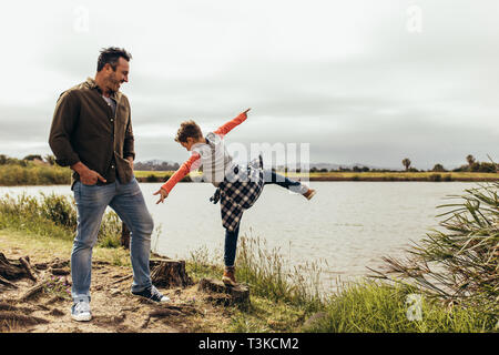 Father and son having fun spending time together playing near the lake. Boy standing on a cut tree trunk on one leg and balancing. Stock Photo