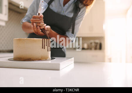 Woman in apron decorating the cake with liquid chocolate. Pastry chef in kitchen decorating cake with chocolate frosting. Stock Photo