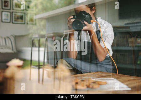 Female inside a kitchen taking pictures of pastries on table with dslr camera. Female chef wearing apron taking pictures of fresh baked dishes. Stock Photo