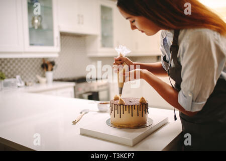 Woman pastry chef decorating chocolate cake in the kitchen. Female wearing a apron decorating cake with a pastry bag with cream. Stock Photo