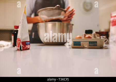 Baking ingredients on kitchen counter with woman sifting flour in the bowl. Pastry chef preparing cake in kitchen. Stock Photo