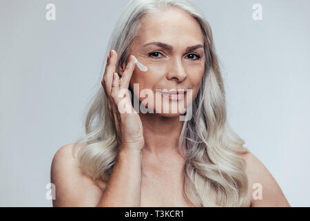 Confident senior woman applying anti aging cream on her face. Beautiful female woman applying moisturizer on her face against grey background. Stock Photo