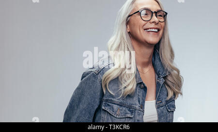 Cheerful senior woman in casuals looking away and smiling over grey background. Happy mature woman in denim shirt and eyeglasses. Stock Photo