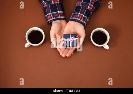 The male hands are closed on a brown background. The guy gives a gift to a girl. Men's hands next to two cups of coffee and with gift. Stock Photo
