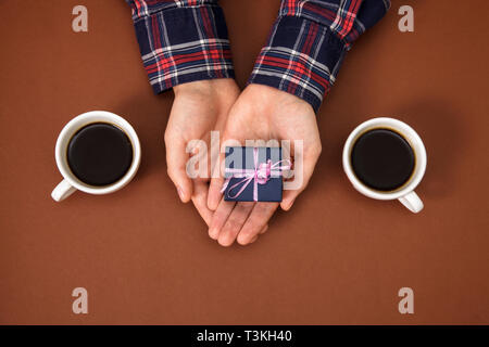 The male hands are closed on a brown background. The guy gives a gift to a girl. Men's hands next to two cups of coffee and with gift. Stock Photo