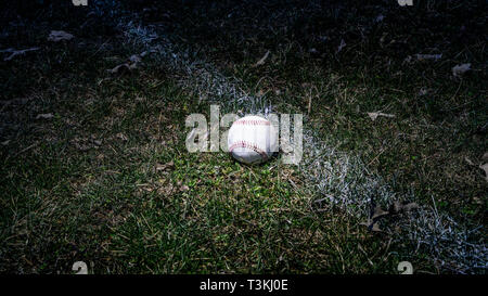 Single Baseball in outfield grass visibly next to chalk line in low light with light shining on Baseball. Stock Photo