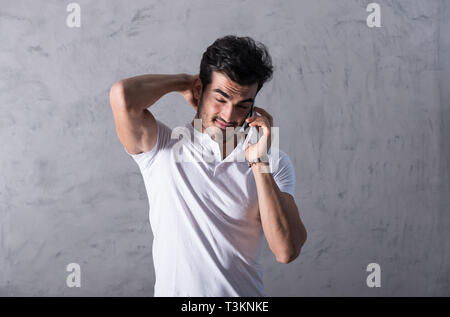 A thinking handsome young man in a white tshirt talking on his phone in front of a grey wall in a studio. Stock Photo