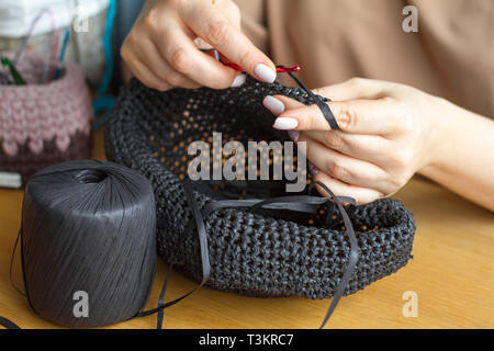 Hands of a young woman knit hat from black synthetic yarn on a light wooden surface. Selective soft focus. Stock Photo