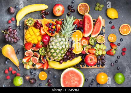 Healthy raw rainbow fruit platter background, mango papaya strawberries oranges blueberries pineapple watermelon on wooden board on dark concrete background, top view, selective focus Stock Photo