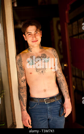Portrait of heavily tattooed young man standing in the doorway of a tattoo parlour. Stock Photo