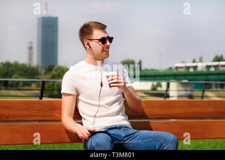 Handsome boy wearing sunglasses using tablet and headphones, sitting on a bench and drinking coffee to go. Handsome student immersed in social media. Stock Photo